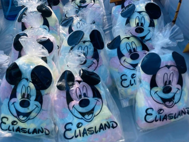 May include: A collection of clear plastic bags filled with colorful cotton candy. Each bag features a smiling Mickey Mouse face and the name "Eliasland" in black text. The cotton candy has pastel colors.