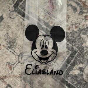 May include: Clear cellophane bag featuring a black Mickey Mouse face and ears. Below the image is the word "Eliasland" in a black, cursive font. The bag is empty and appears to be for party favors or small gifts.
