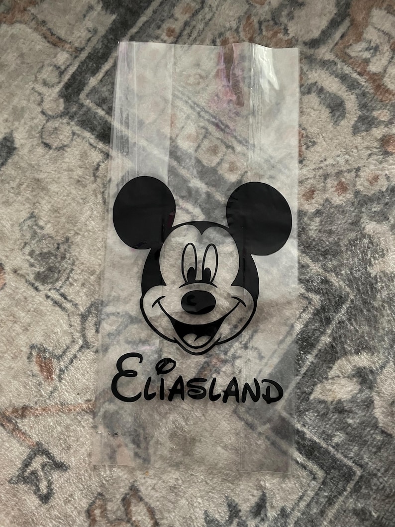 May include: Clear cellophane bag featuring a black Mickey Mouse face and ears. Below the image is the word "Eliasland" in a black, cursive font. The bag is empty and appears to be for party favors or small gifts.