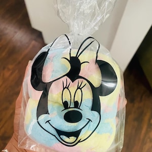 May include: A clear plastic bag featuring a black Minnie Mouse design, filled with colorful, pastel-colored candy. The candy appears to be a mix of blue, yellow, pink, and white. The bag is tied at the top.