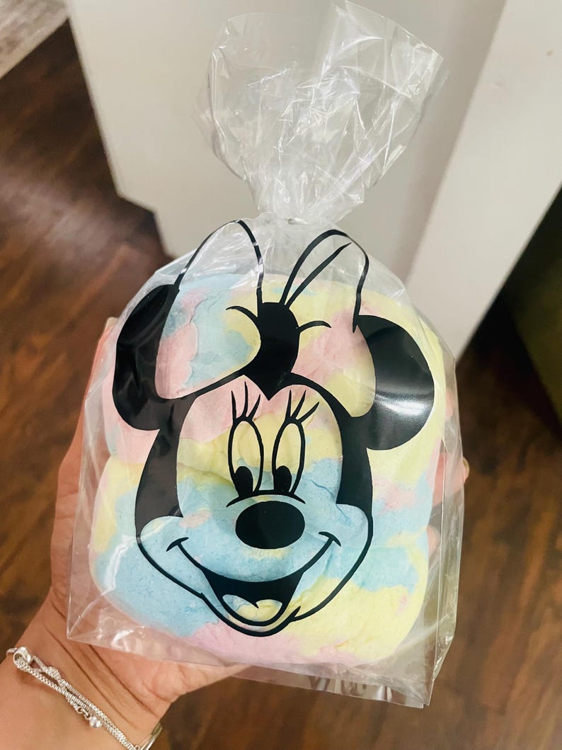 May include: A clear plastic bag featuring a black Minnie Mouse design, filled with colorful, pastel-colored candy. The candy appears to be a mix of blue, yellow, pink, and white. The bag is tied at the top.