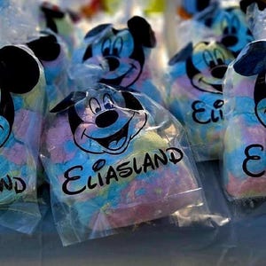 May include: Several clear plastic bags filled with colorful cotton candy. Each bag features a Mickey Mouse design and the text "Eliasland". The cotton candy is a mix of blue, pink, and yellow.