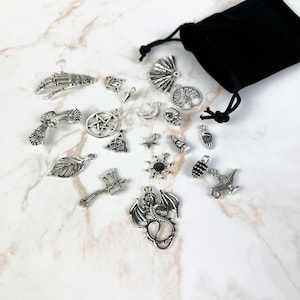Op de afbeelding: Een verzameling zilveren bedelarmbanden in een zwarte fluwelen zak. De bedels omvatten een draak, een levensboom, een pentagram, een hand, een zon, een halve maan, een blad, een bijl, een schedel, een ster, een lamp, een dennenappel, een waaier, een vogel, een skelethand, een knoop en een wenslamp.