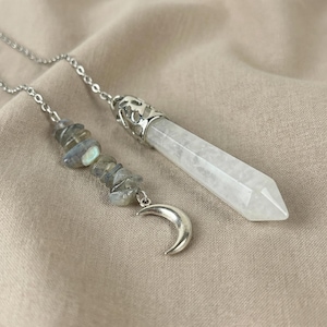 Può includere: Una collana a catena color argento con un ciondolo a punta di cristallo trasparente sfaccettato e un cappuccio decorativo in metallo. Un segmento di catena secondario mostra una serie di piccole pietre irregolari grigio-blu e un ciondolo a mezzaluna.