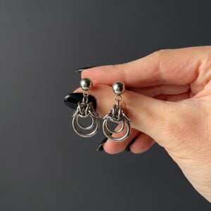 Puede incluir: Un par de pendientes plateados. Cada pendiente presenta una pequeña bola en la parte superior, conectada a un elemento decorativo con dos anillos colgantes. Los pendientes se muestran sobre un fondo gris oscuro.