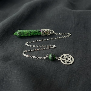 Puede incluir: Un péndulo verde y plateado con un dije de pentagrama. El péndulo presenta un cristal verde facetado con inclusiones verde oscuro, conectado por una cadena plateada a una pequeña piedra verde y un dije de pentagrama. El fondo es una tela gris oscura.