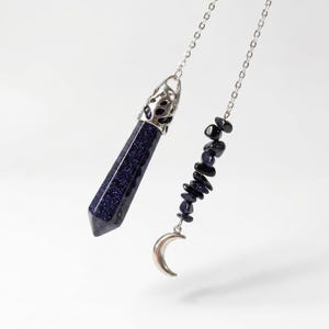 May include: A silver-toned chain necklace featuring a dark blue glittery sandstone pendant in a faceted hexagonal point shape with a decorative metal cap, alongside a smaller strand of dark blue stone chips ending in a silver crescent moon charm.
