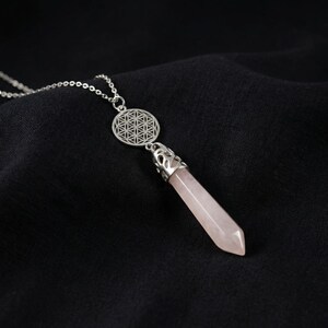 May include: A silver-toned necklace featuring a pale pink, hexagonal-cut rose quartz crystal pendant capped with an ornate metal bail. Above the crystal hangs a circular silver charm displaying an intricate, geometric flower of life pattern.