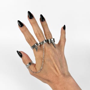 Puede incluir: Anillos plateados unidos por una cadena, cada uno con un diseño puntiagudo. Los anillos se usan en los dedos índice, medio, anular y pulgar. Las uñas están pintadas de negro.