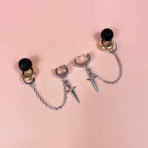 Puede incluir: Pendientes plateados con un dije de daga y una cadena que conecta una cuenta negra y un anillo dorado. Los pendientes se muestran sobre un fondo rosa claro.