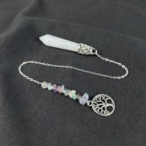 Könnte beinhalten: Ein weißes Kristallpendel mit einer Silberkette und einem Lebensbaum-Anhänger. Die Kette hat auch eine Reihe von bunten, kleinen, rohen Edelsteinen. Das Pendel ist für Wahrsagerei und spirituelle Praktiken konzipiert.