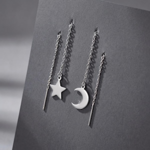 Puede incluir: Pendientes colgantes plateados con un dije de estrella y media luna. Los dijes cuelgan de delicadas cadenas, con una barra recta al final de cada cadena. Los pendientes se exhiben sobre un fondo gris oscuro.