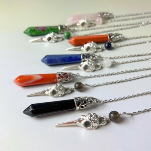 Könnte beinhalten: Eine Sammlung von sechs Halsketten mit hexagonalen Kristallanhängern in verschiedenen Farben wie Grün, Orange, Blau und Schwarz. Jeder Anhänger ist mit einem silberfarbenen Metall-Vogelschädel-Anhänger und einer dünnen Silberkette verbunden.