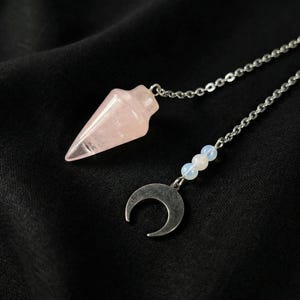 Puede incluir: Un péndulo de cuarzo rosa facetado de color rosa claro unido a una cadena de metal plateado. La cadena presenta un pequeño dije de media luna de plata oscura acentuado por tres diminutas cuentas redondas y translúcidas en tonos azul pálido y blanco.