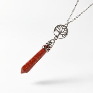 May include: A necklace featuring a hexagonal, glittery, reddish-brown goldstone pendant capped with a silver-toned filigree metal piece. Above the pendant is a circular silver-toned charm depicting a tree with spreading branches and roots.