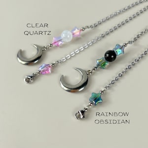 May include: Three silver chain necklaces with crescent moon pendants. Each necklace features a different gemstone: clear quartz, a black stone, and rainbow obsidian. The necklaces also have iridescent star-shaped beads. The text "CLEAR QUARTZ" and "RAINBOW OBSIDIAN" are visible.