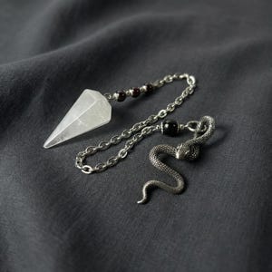 Op de afbeelding: Een kristallen slinger met een facetgeslepen, heldere kwartspunt, verbonden door een zilveren ketting met rode kralen en een zwarte kraal. Er is ook een zilveren slangensieraad bevestigd. De items liggen op een donkergrijze stof.