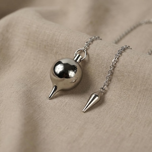 May include: A polished silver-toned metal pendulum with a spherical top and a pointed base, suspended from a matching silver-toned metal chain. The chain features a smaller, slender, pointed metal weight at the end, creating a dual-component design.