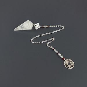 May include: A clear, faceted crystal pendulum attached to a silver-toned metal chain. The chain features a decorative floral-shaped metal charm, small dark red beads, and clear beads, ending in a circular metal charm with a geometric pattern.