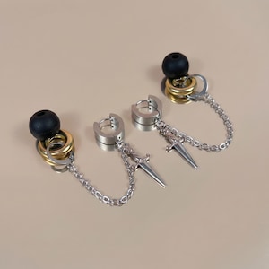 Puede incluir: Pendientes de aro plateados con un pequeño dije de daga colgando de una cadena. Cada pendiente tiene una bola negra y anillos dorados y plateados unidos en la parte superior. Los pendientes se exhiben sobre un fondo neutro.