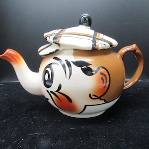 May include: A ceramic teapot shaped like a character head with a tan face, black painted eyes and hair, and a red-tipped spout. The lid is designed as a white, brown, and black plaid flat cap with a small black knob handle on top.
