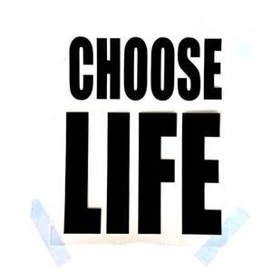 May include: A white paper with the words "CHOOSE LIFE" in bold, black capital letters. The paper is secured to a white surface with two pieces of blue tape.