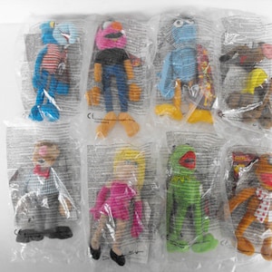 May include: A collection of eight Muppet plush toys individually sealed in clear plastic packaging. The set includes characters with various colors such as blue, pink, green, and brown, featuring distinct outfits like striped shirts, dresses, and bow ties.