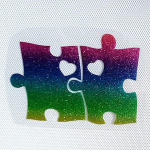 Puede incluir: Dos piezas de rompecabezas de purpurina arcoíris encajan para formar una forma de corazón. Las piezas del rompecabezas tienen corazones de purpurina blanca en el centro.