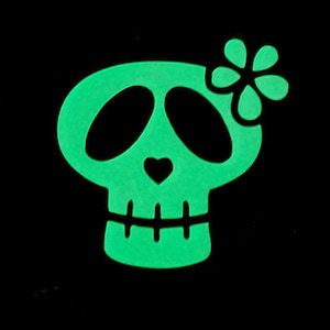 May include: A green glow-in-the-dark skull with a flower on top. The skull has a heart shape on its forehead and a toothy grin.