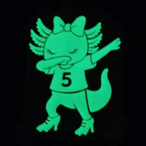 Può includere: Una toppa grafica luminescente che raffigura un axolotl dei cartoni animati in posa dab. L'axolotl indossa un fiocco sulla testa, una maglietta con il numero 5 stampato sul davanti e scarpe con i tacchi alti. Il design è verde brillante su sfondo nero.