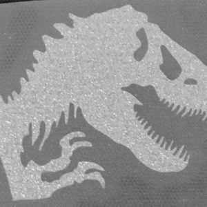 May include: A white silhouette of a dinosaur, possibly a Tyrannosaurus Rex, with a large head and sharp teeth. The dinosaur is facing right and has a textured, glittery appearance.
