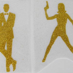 Op de afbeelding: Twee gouden glitter silhouetten op een witte achtergrond. Eén figuur is een man in smoking met een pistool. De andere is een vrouw met een pistool, in een dynamische pose. De figuren zijn gedetailleerd met glitter.