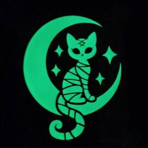 Peut inclure: Un motif graphique phosphorescent sur fond noir représentant un chat momie enveloppé de bandelettes assis sur un croissant de lune. Le design comprend des étoiles à quatre branches entourant le chat, le tout en vert lumineux éclatant.
