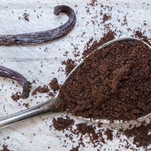 Puede incluir: Primer plano de una cuchara de metal llena de granos de vainilla molidos. Los granos son de color marrón oscuro y tienen una textura fina. También hay dos vainas de vainilla enteras en el fondo.