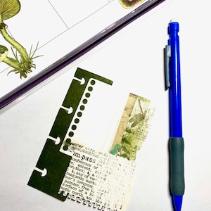 Op de afbeelding: Een blauw mechanisch potlood met een grijze grip ligt naast een donkergroen papier met een witte binnenkant en een gescheurd stuk papier met tekst. Een notitieboek met paddenstoelillustraties staat op de achtergrond.