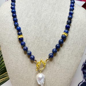 May include: A beaded necklace featuring deep blue spherical beads and gold-colored accents. The necklace has a gold clasp and a large, white, irregularly shaped pearl pendant. The necklace is displayed on a neutral-toned surface.