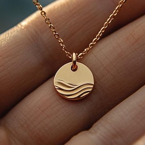 Pode incluir: Um colar dourado com uma corrente delicada e um pingente redondo. O pingente apresenta um design de ondas. O colar é exibido em uma mão, mostrando o tamanho e os detalhes da joia.