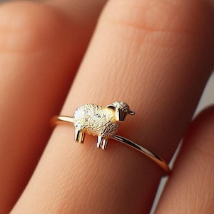 May include: A gold ring with a small, detailed sheep charm. The ring is worn on a finger.