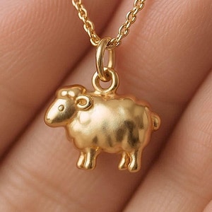 May include: A gold necklace with a sheep pendant. The pendant is a detailed, three-dimensional sheep figure with a textured surface. The chain is a delicate, linked design. The necklace is shown against a soft background.