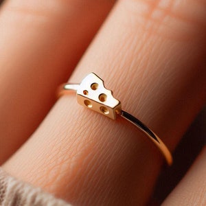 Puede incluir: Un anillo de oro con un pequeño dije de queso detallado. El dije de queso tiene agujeros y tiene forma de cuña.