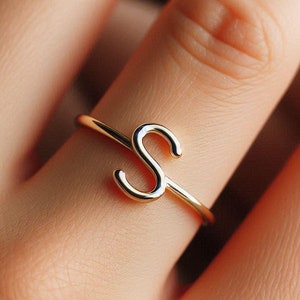 Op de afbeelding: Een ring van goudkleur met een simpel, cursief letter 'S' ontwerp. De ring wordt aan een vinger gedragen.