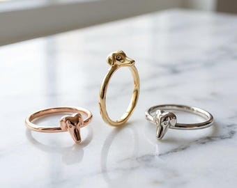 May include: Three metal rings, each featuring a small sculpted dog head as the focal point. One ring is rose gold, one is yellow gold, and one is silver-toned. Each band is thin and circular, showcasing a polished finish on the metal surfaces.