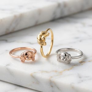 May include: Three rings featuring a sculpted dog head with long, floppy ears. One ring is rose gold, one is yellow gold, and one is silver. Each band is thin and circular, showcasing the detailed metalwork of the canine face design.