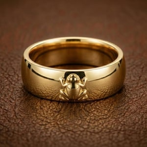 May include: A gold-colored ring with a frog design. The ring is made of a shiny, polished metal and features a detailed frog emblem on the front. The ring is set against a brown leather-textured surface.