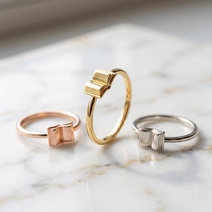 May include: Three thin metal rings, each featuring a small, open book charm at the center. The set includes one rose gold ring, one yellow gold ring, and one silver-toned ring, all displayed with a smooth, polished finish and simple bands.