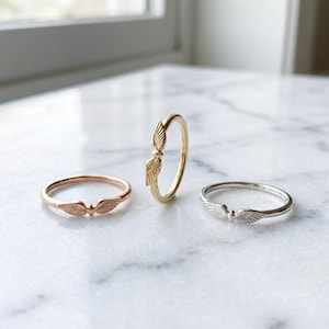 May include: Three thin metal rings, each featuring a small pair of engraved wings at the center. One ring is rose gold, one is yellow gold, and one is silver. The wings on each ring are detailed with fine lines to mimic the appearance of feathers.