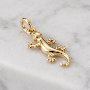 Puede incluir: Un colgante de metal dorado pulido con forma de pequeña lagartija o gecko. El dije presenta una superficie lisa y reflectante con dedos detallados y una cola curva, e incluye una argolla circular en la parte superior para una cadena.