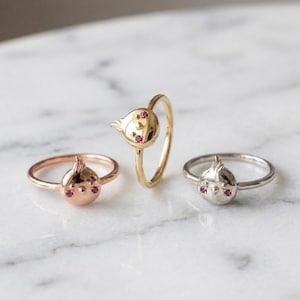 May include: Three rings featuring a bird-shaped design with two small pink gemstone eyes. The rings are crafted in three different metal finishes: rose gold, yellow gold, and silver. Each bird motif includes a small crest detail on top.