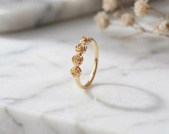 Può includere: Un anello in oro con una fascia decorata da tre piccoli motivi a rosa scolpiti, disposti in fila verticale sulla parte superiore. Il metallo presenta una finitura lucida e riflettente, con dettagli intricati dei petali su ogni rosa.