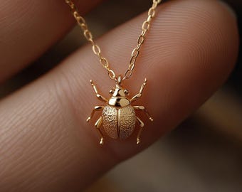 Könnte beinhalten: Eine goldfarbene Halskette mit einem detaillierten Käferanhänger. Der Anhänger hat strukturierte Flügel und zarte Beine, die an einer feinen Kette hängen. Der Schmuck wird vor einem unscharfen Hintergrund präsentiert.
